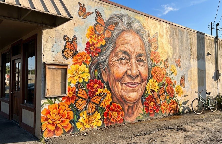 Large portrait mural of a smiling older woman surrounded by marigolds and monarch butterflies on a restaurant wall