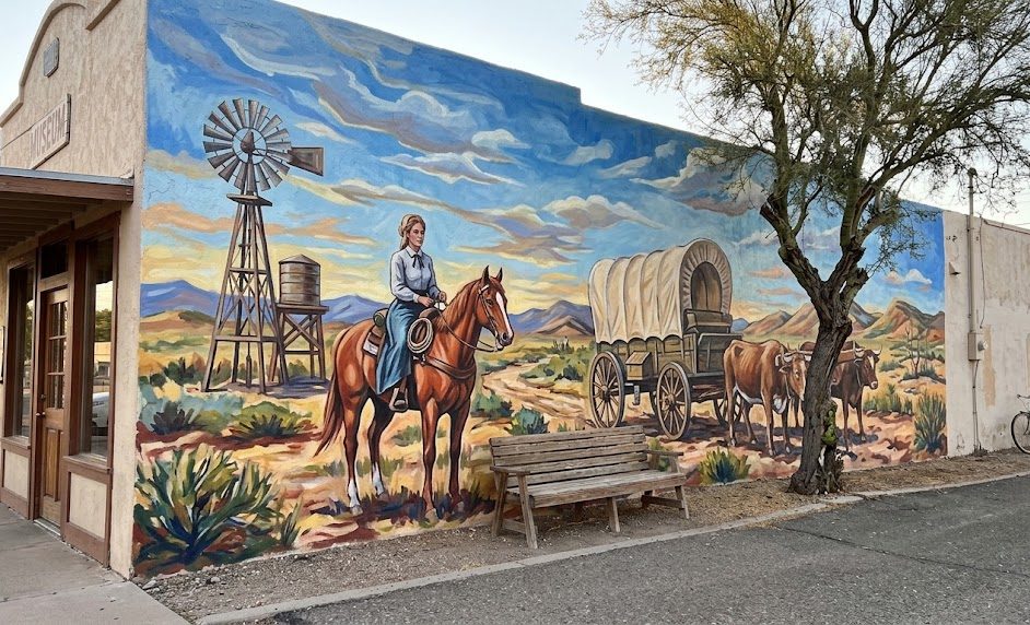 Mural of ranch life with a woman on horseback, a covered wagon and a windmill on a long stucco wall